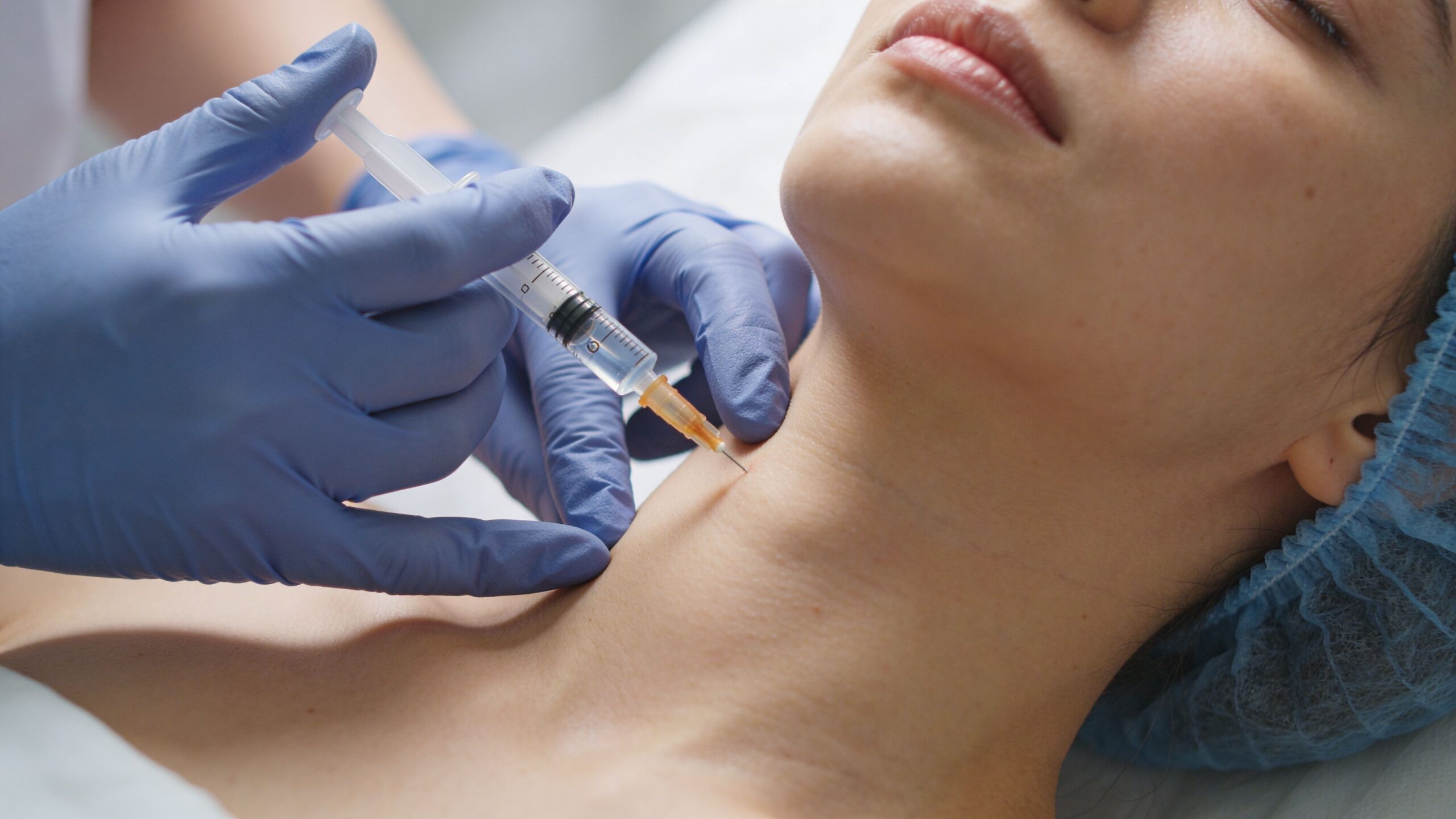A dermatologist administers a cosmetic neck injection to a patient lying down in a medical clinic office.