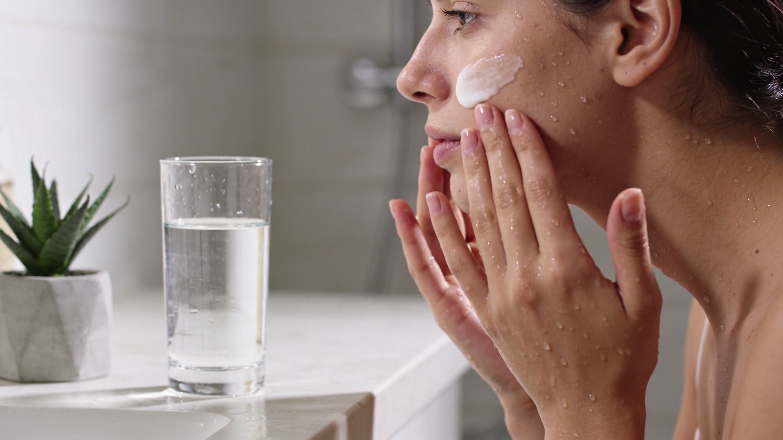 A young woman applying white skincare cream to her wet face while standing near a glass of water.