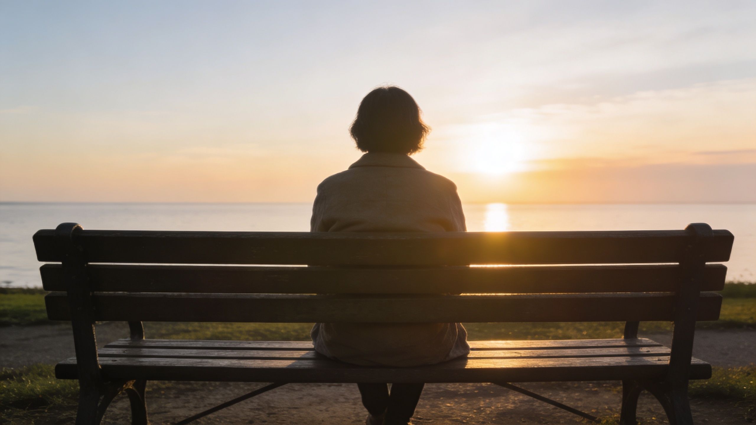 A person sitting alone on a park bench watching the sunset over the horizon of the ocean.